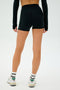 A person stands against a plain backdrop, wearing the SPLITS59 Ella Airweight High Waist Short 3.5" in Black/Arugula alongside a long-sleeve black top, white socks, and sneakers, with their back facing the camera.