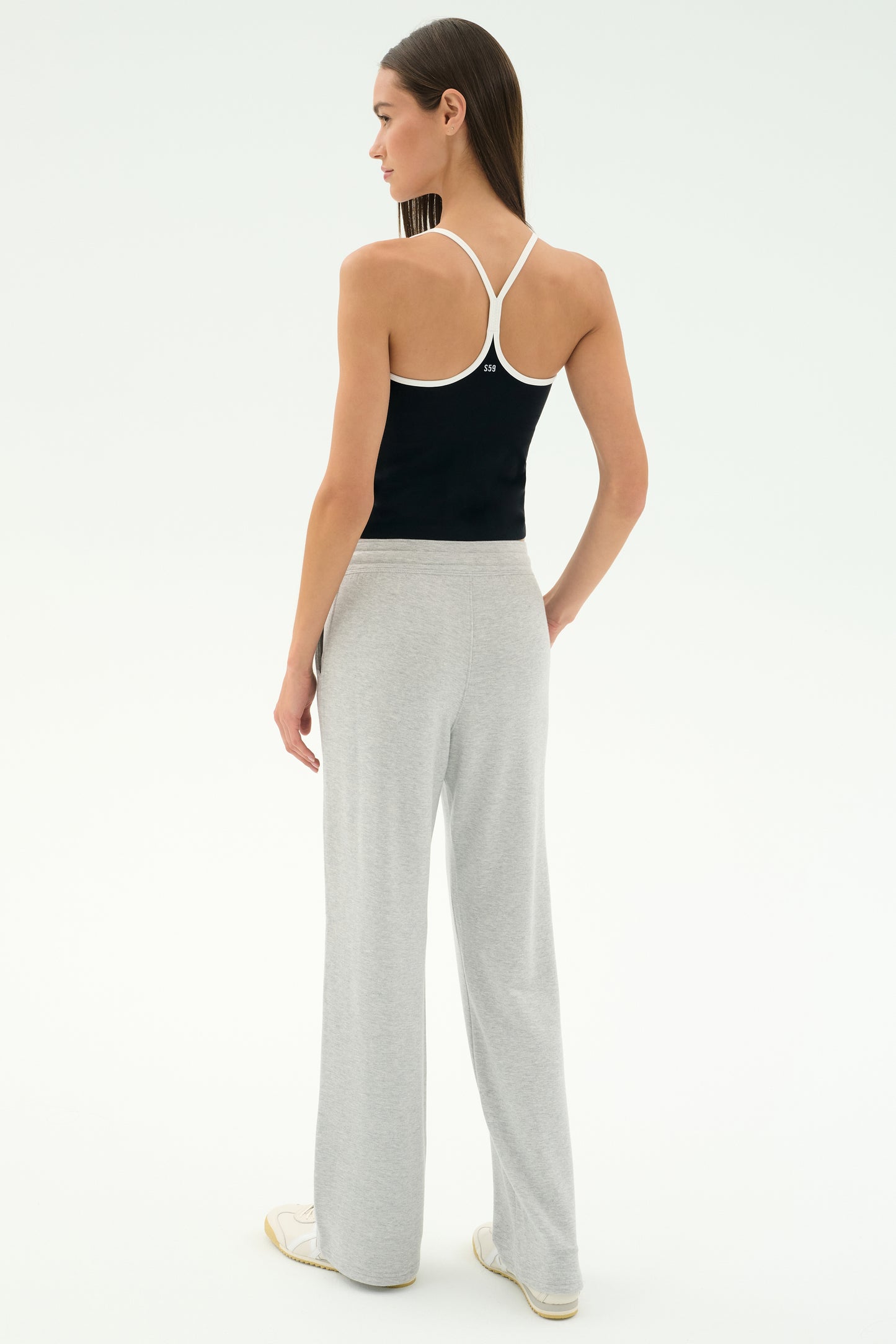 A woman stands facing away, wearing a black racerback tank top, SPLITS59 Raven Fleece Full Length Sweatpant in Heather Grey, and white sneakers against a plain background.