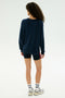 Someone with curly hair stands facing away on a plain light background, wearing the SPLITS59 Andie Fleece Sweatshirt in Indigo, paired with black shorts, white socks, and sneakers.