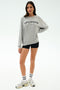 A woman faces forward in the SPLITS59 Andie Oversized Fleece Varsity Graphic Sweatshirt in heather grey, paired with black shorts, white socks, and sneakers against a plain white background.