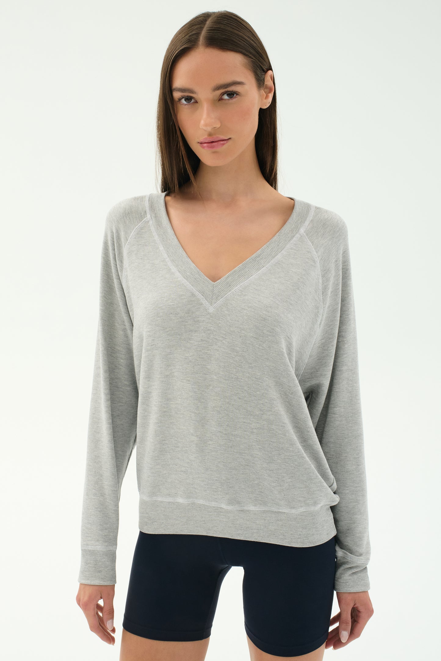 A woman stands against a plain white background in the SPLITS59 Bennie Fleece V Neck Sweatshirt in Heather Grey/White, made in Los Angeles, paired with black shorts for a chic, casual look.