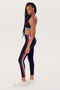 A woman with long braids is posing in athletic wear made from nylon and spandex fabric, featuring a black sports bra with red accents (Bianca Rigor Bra - Black by SPLITS59) and black leggings adorned with red and white stripes. She completes the look with white sneakers.