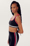 A person with long braids wearing a black SPLITS59 Bianca Rigor Bra - Black and black leggings made from spandex with white and red stripes stands against a plain background.