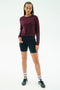 A person stands against a white background wearing the SPLITS59 Brooks Fleece Long Sleeve Sweatshirt in Dark Cherry, paired with black cycling shorts, white striped socks, and dark sneakers.