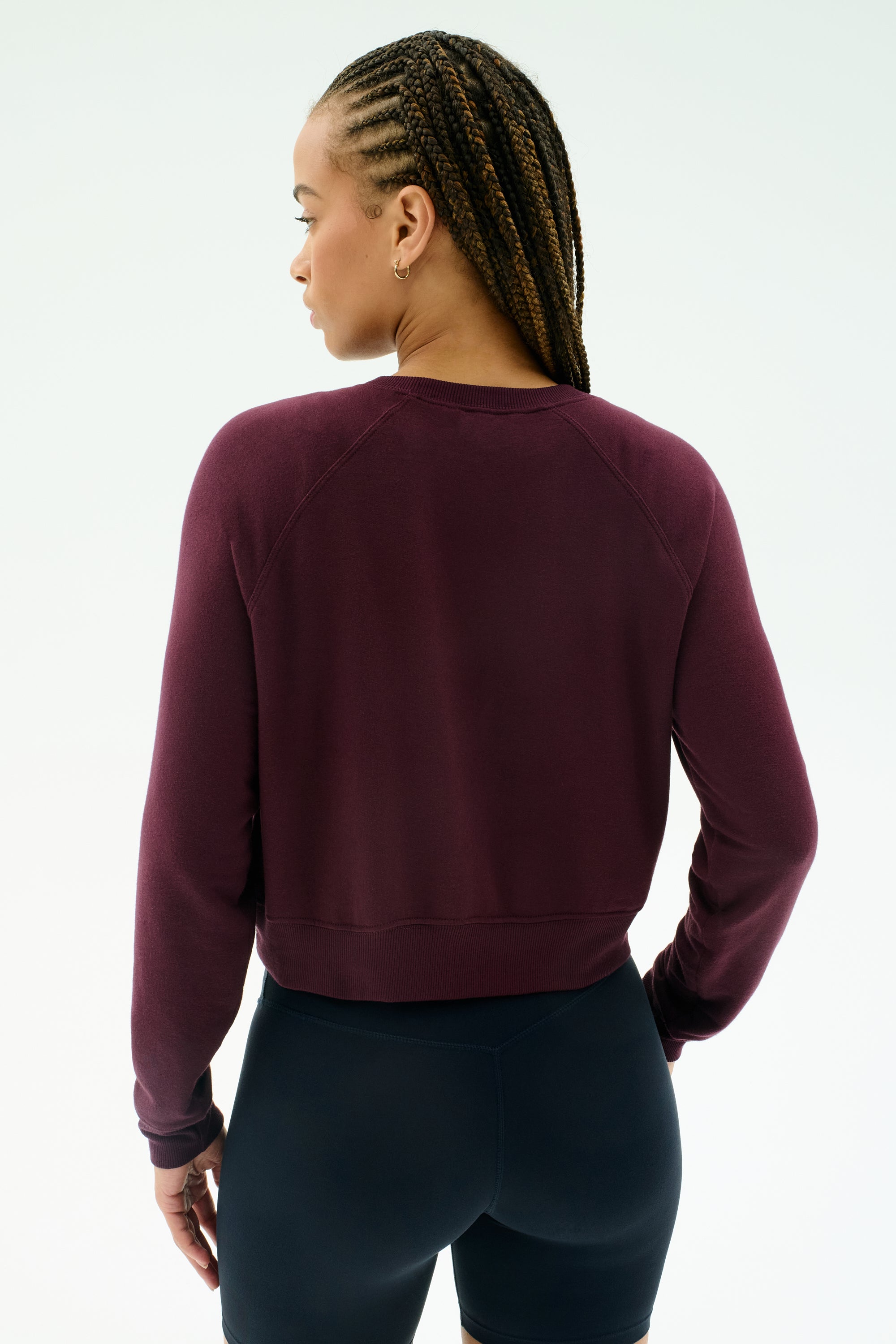 A person with braided hair wears the SPLITS59 Brooks Fleece Long Sleeve Sweatshirt in Dark Cherry and black fitted modal spandex shorts, facing away against a plain light background.