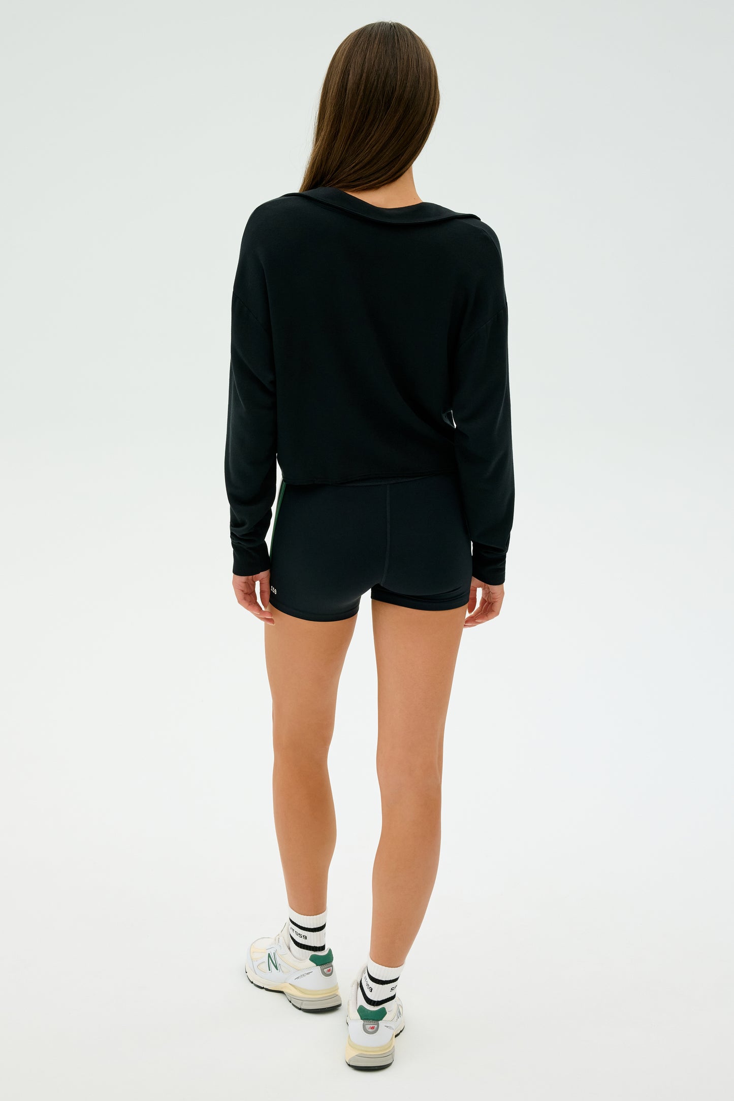 A woman stands facing away, wearing a Joan Fleece Polo Sweatshirt in black by SPLITS59, styled with shorts, white sneakers, and striped socks against a plain background.
