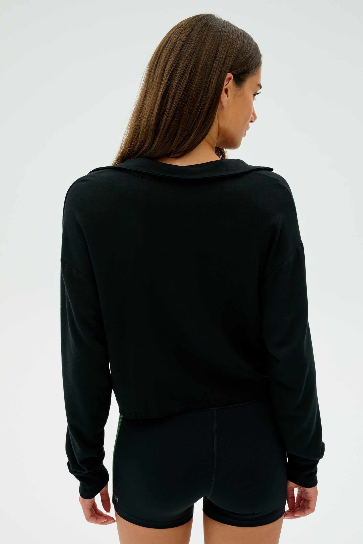 A long-haired individual is facing away, wearing SPLITS59's Joan Fleece Polo Sweatshirt in black along with black shorts.