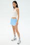 A woman stands against a plain white background wearing the SPLITS59 Megan Woven Nylon Run Short in Skylight, a white spandex tank top, striped socks, and sneakers.