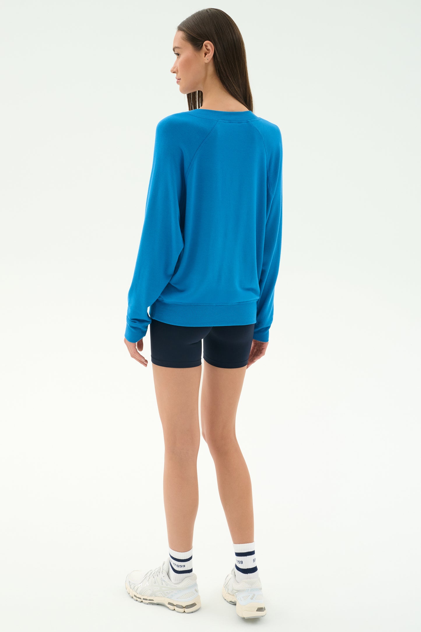A woman faces away, wearing the SPLITS59 Bennie Fleece V-Neck Sweatshirt in Stone Blue with navy shorts, white striped socks, and white sneakers against a plain light background.