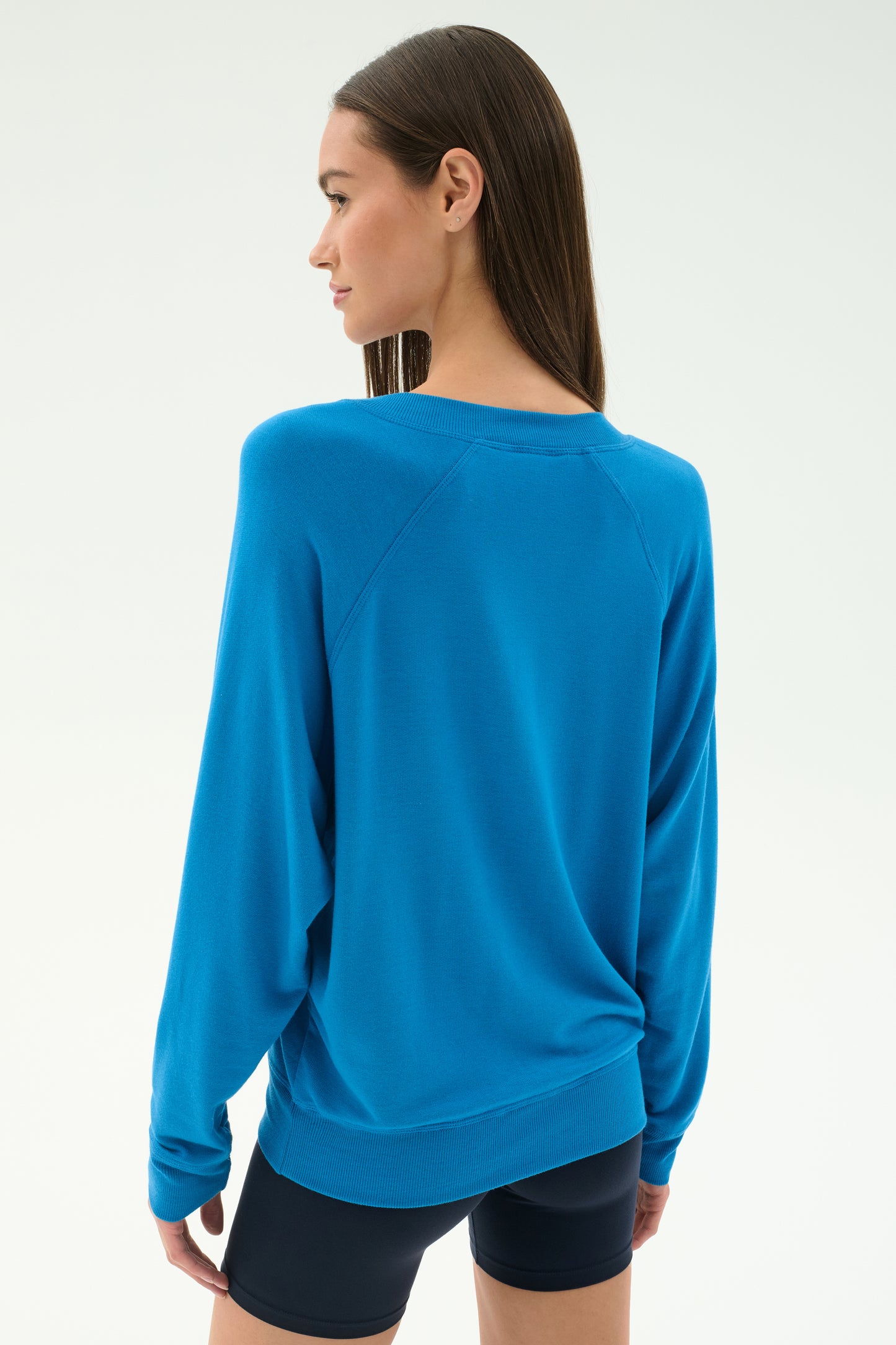Woman wears the SPLITS59 Bennie Fleece V-Neck Sweatshirt in Stone Blue with black shorts, facing away from the camera against a plain light background.