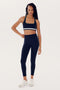 A woman with long braided hair poses in a Bianca Rigor Bra - Indigo by SPLITS59 and matching high-waisted leggings made from stretchy spandex fabric. She is wearing white sneakers and standing against a plain white background.