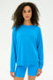 A woman with curly hair wears the SPLITS59 Andie Fleece Sweatshirt in Riviera— a loose, blue crewneck pullover made from soft stretch modal—paired with matching blue shorts, standing against a plain white background.
