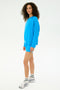 A person with curly hair wears the SPLITS59 Andie Fleece Sweatshirt - Riviera and matching shorts, along with white socks and sneakers, posing against a plain white background. MADE IN LOS ANGELES.
