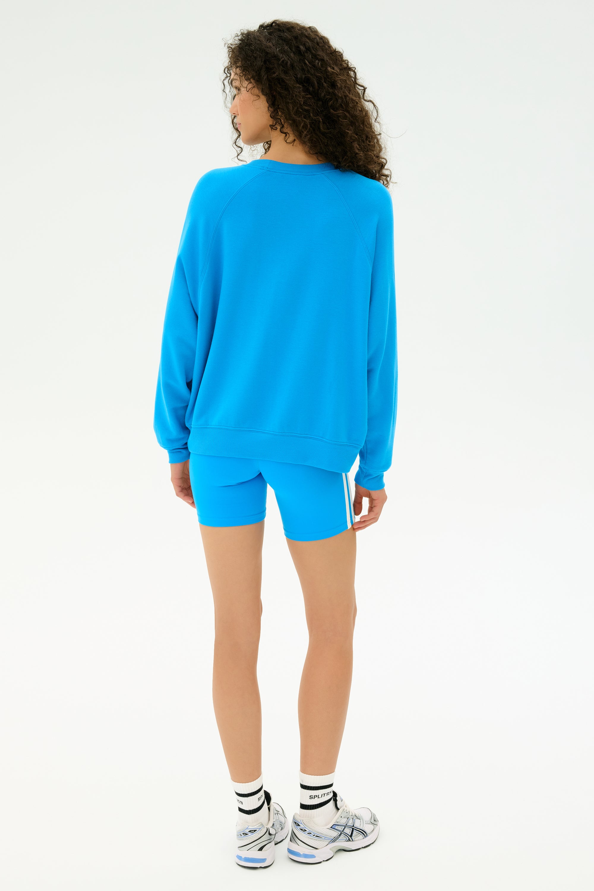 A person faces away, wearing the SPLITS59 Andie Fleece Sweatshirt in Riviera blue with matching shorts, white socks, and silver sneakers against a plain white background. MADE IN LOS ANGELES.