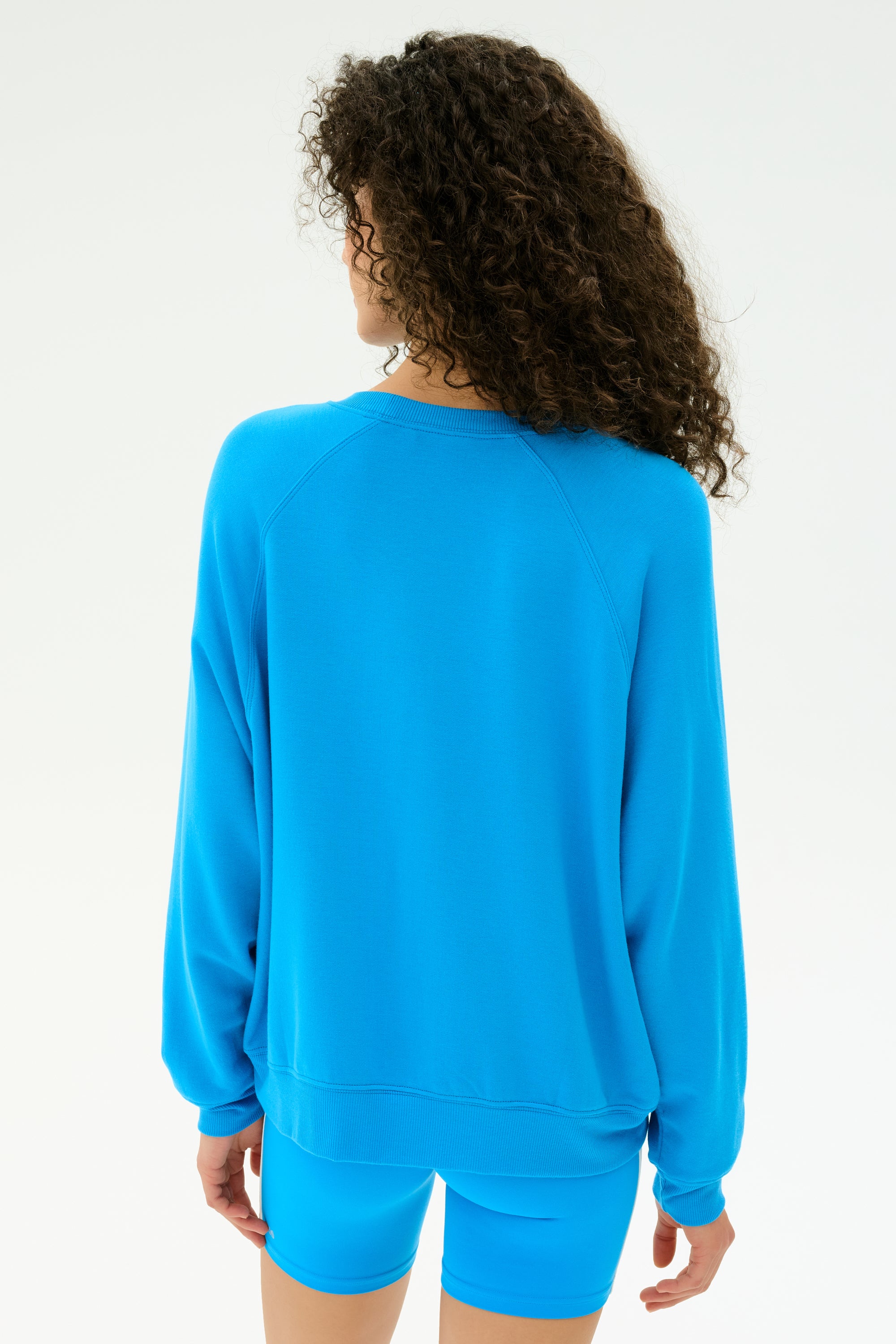 A person with curly hair, facing away, wears the SPLITS59 Andie Fleece Sweatshirt in Riviera— a bright blue crewneck pullover made from stretch modal fleece—set against a plain light background. Made in Los Angeles.