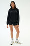 A woman faces forward in a black SPLITS59 Andie Oversized Fleece Varsity Graphic Sweatshirt with "Los Angeles" text, black shorts, white socks, and sneakers against a plain white background. Made in Los Angeles.