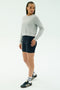 A person stands slightly to the side wearing the SPLITS59 Brooks Fleece Long Sleeve Sweatshirt in Heather Grey, paired with navy cycling shorts, white socks, and black Adidas sneakers against a plain light background.