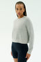 A person with braided hair wears the SPLITS59 Brooks Fleece Long Sleeve Sweatshirt in Heather Grey and black fitted spandex-blend shorts, standing against a plain light background.