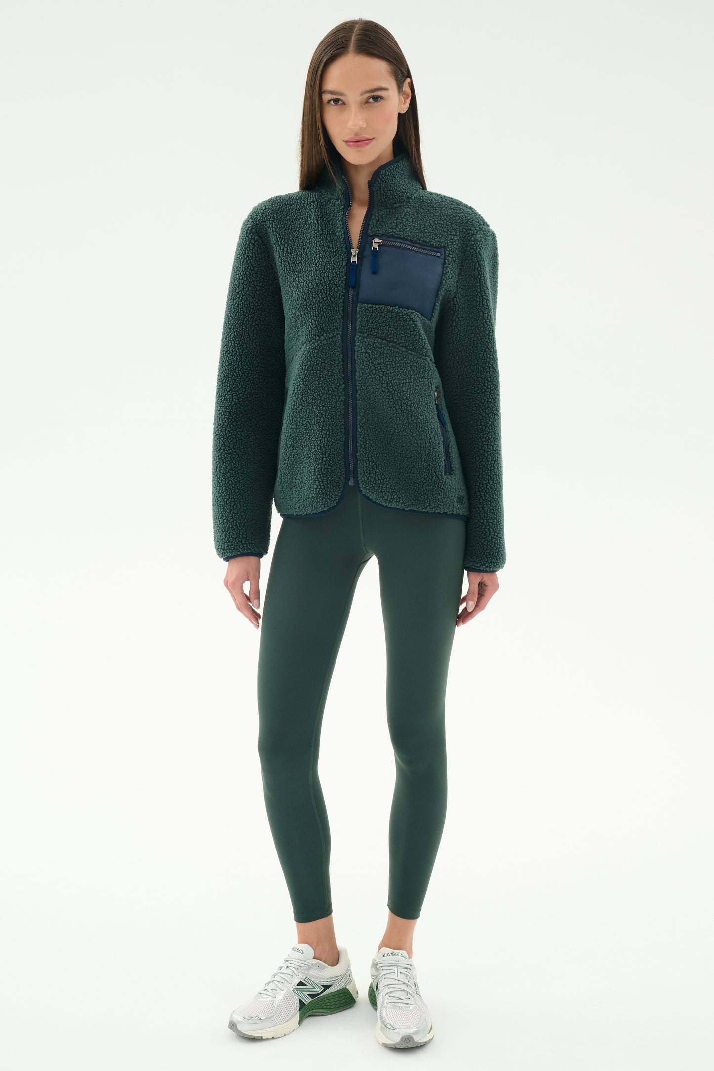A woman wears the SPLITS59 Harris Sherpa Jacket in Military/Indigo, paired with dark green leggings and white sneakers, standing against a plain light background.