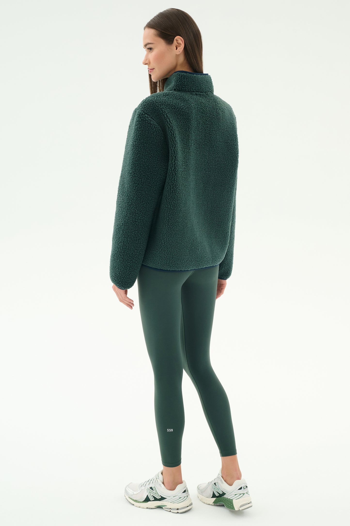 A woman faces away, wearing the SPLITS59 Harris Sherpa Jacket in Military/Indigo, with matching leggings and white sneakers against a plain white background.