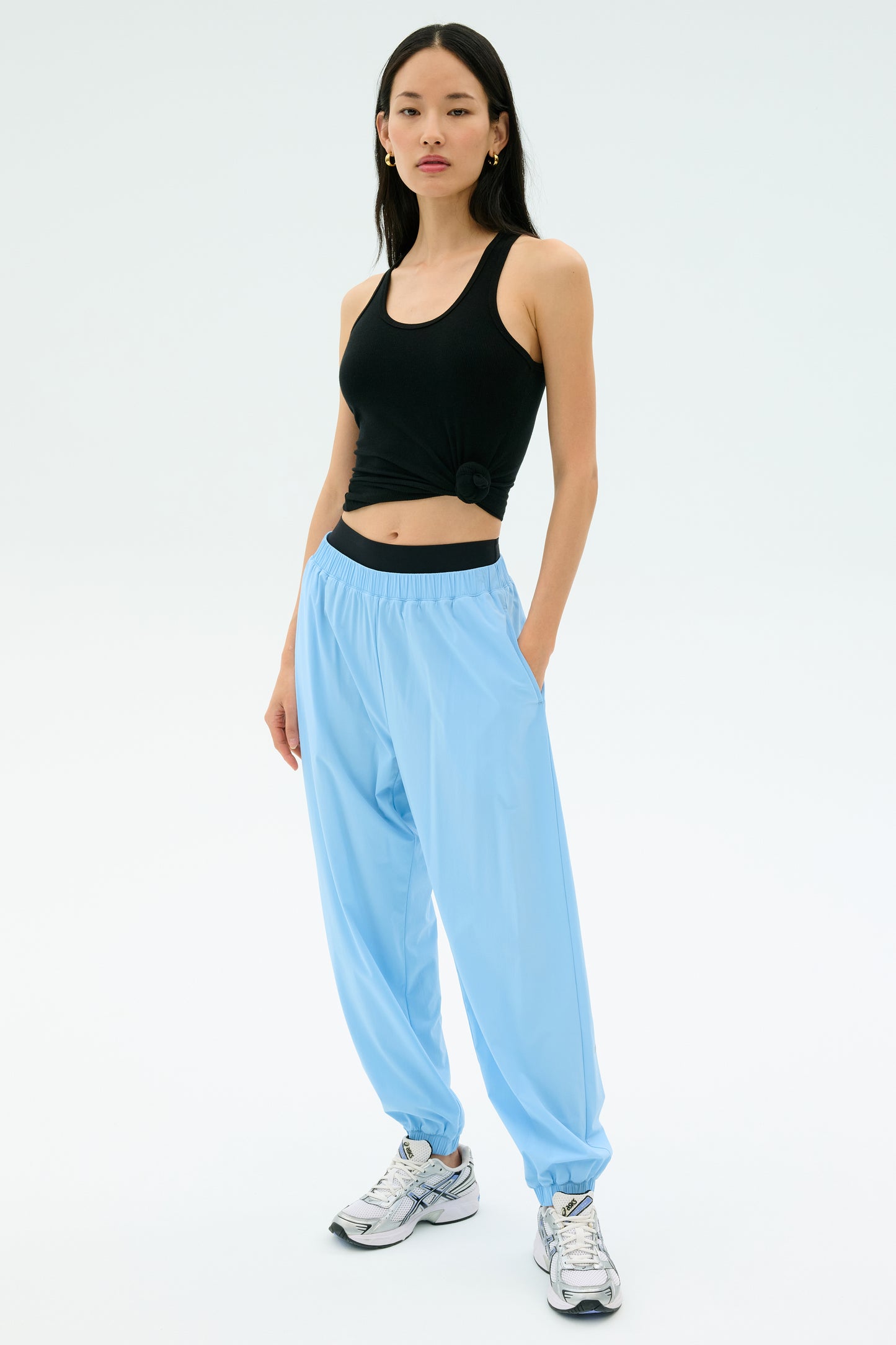 A person wearing a black sleeveless top, SPLITS59 Megan Woven Nylon Jogger in Skylight, and white sneakers stands against a plain light background.
