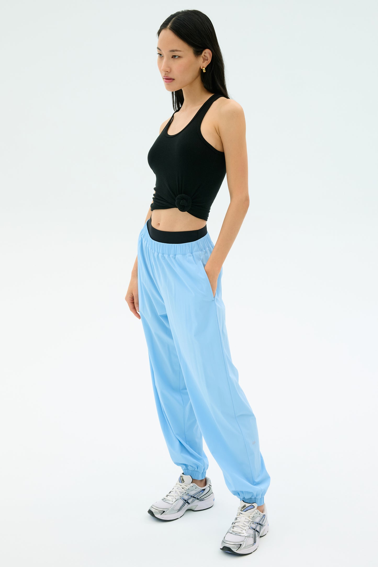 A woman stands against a plain background wearing SPLITS59's Megan Woven Nylon Jogger in Skylight, paired with a black sleeveless crop top and white sneakers, hands in her pockets.