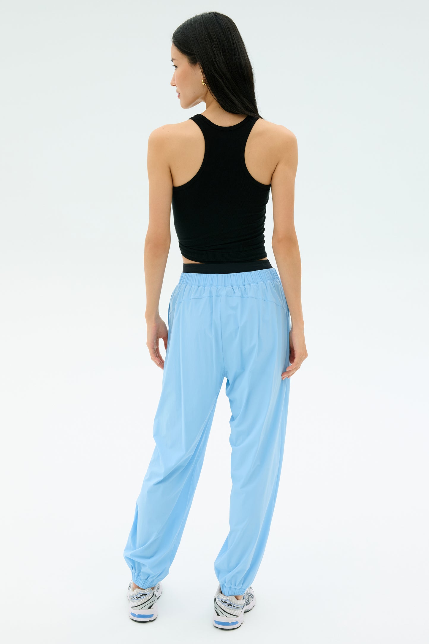A woman faces away, wearing a black sleeveless top, SPLITS59 Megan Woven Nylon Jogger in Skylight, and sneakers against a plain white background.
