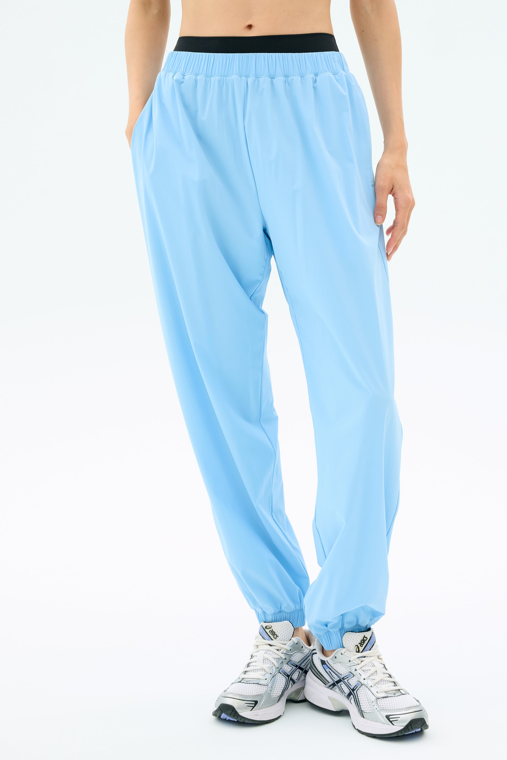 A person wears SPLITS59’s Megan Woven Nylon Jogger in Skylight with white athletic shoes, hands in pockets, standing against a plain white background.