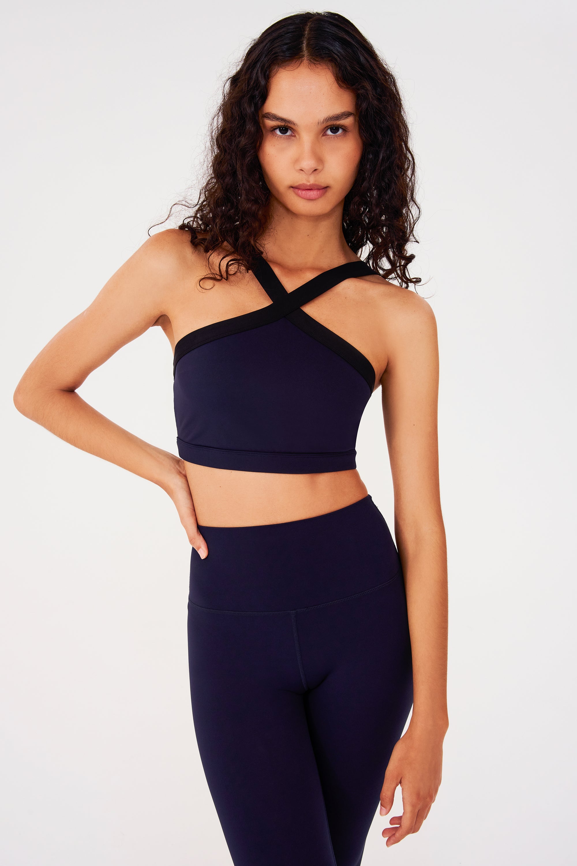 Front view of woman with dark wavy hair wearing a dark blue bra with thick black cross-front neckline straps and dark blue leggings