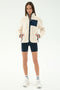 A woman stands facing forward wearing the SPLITS59 Harris Sherpa Jacket in Creme/Indigo, paired with navy shorts, white socks, and white sneakers against a plain light background.