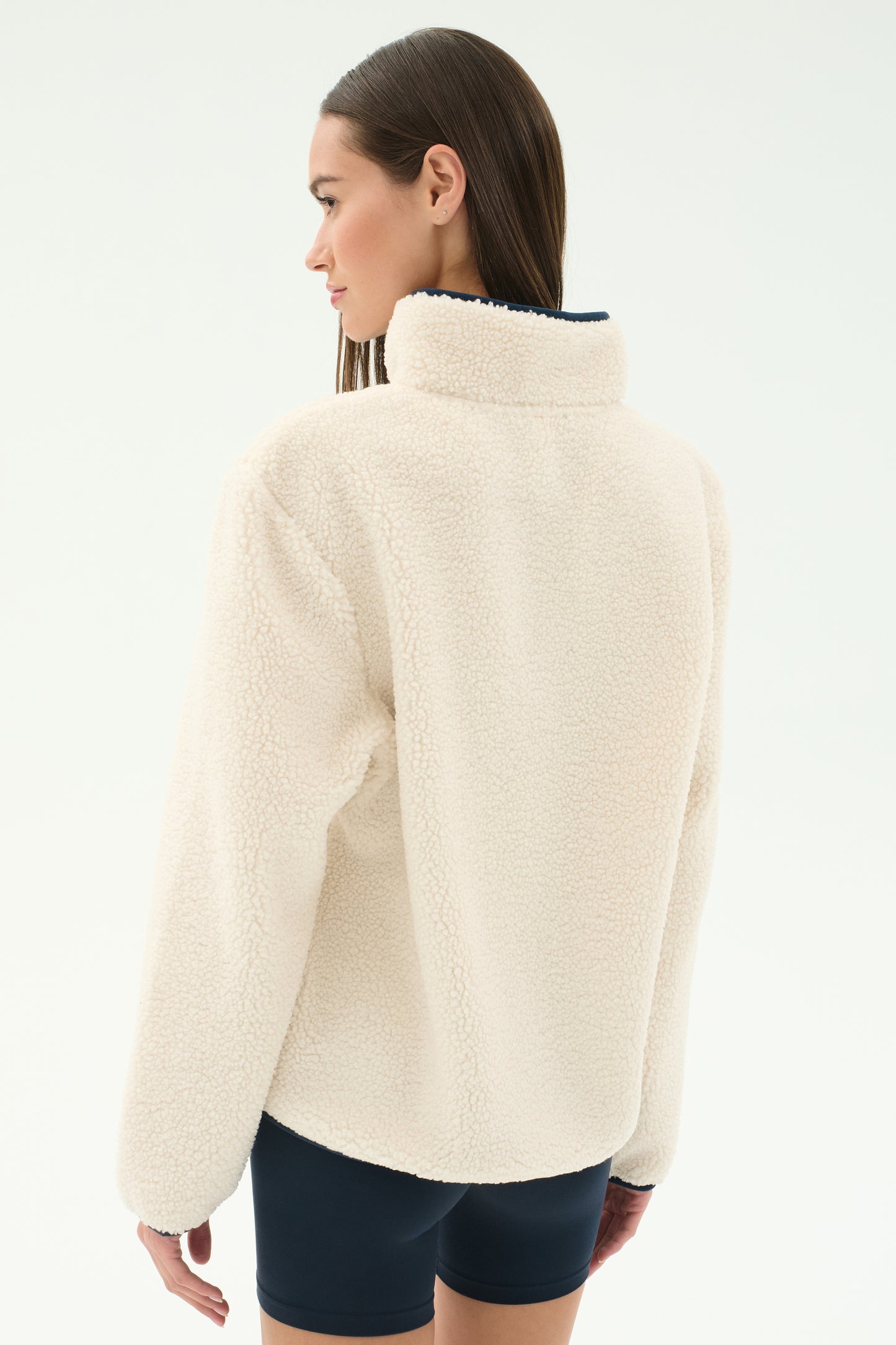 A woman with straight brown hair is seen from the back in a SPLITS59 Harris Sherpa Jacket in Creme/Indigo and dark shorts, against a plain white background.