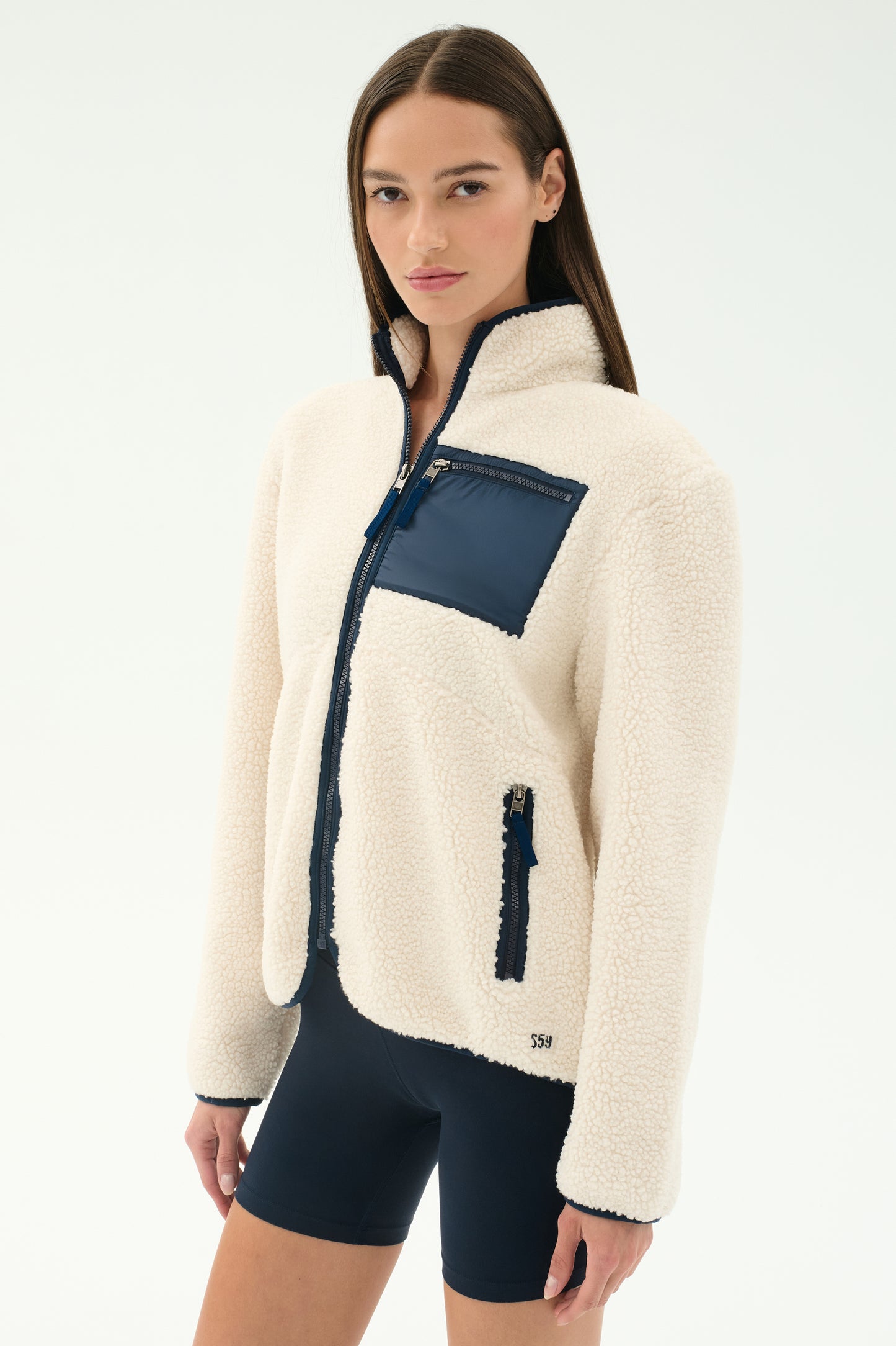 A woman models the SPLITS59 Harris Sherpa Jacket in Creme/Indigo, featuring navy zipper and pocket details, paired with black shorts, against a plain white background.