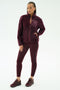 A woman stands against a white background wearing the SPLITS59 Harris Sherpa Jacket in Dark Cherry, paired with matching leggings featuring a black stripe and burgundy sneakers.