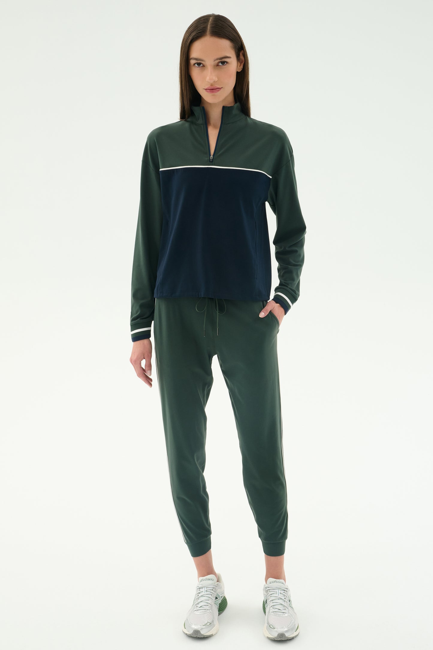A woman faces forward in the SPLITS59 Millie Airweight Half Zip in Military/Indigo, a soft nylon tracksuit with a front zip, paired with white sneakers, against a plain white background.