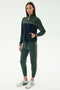 A woman wears the SPLITS59 Millie Airweight Half Zip in Military/Indigo, featuring a spandex-blend with white piping, and white athletic shoes, standing against a plain light background.