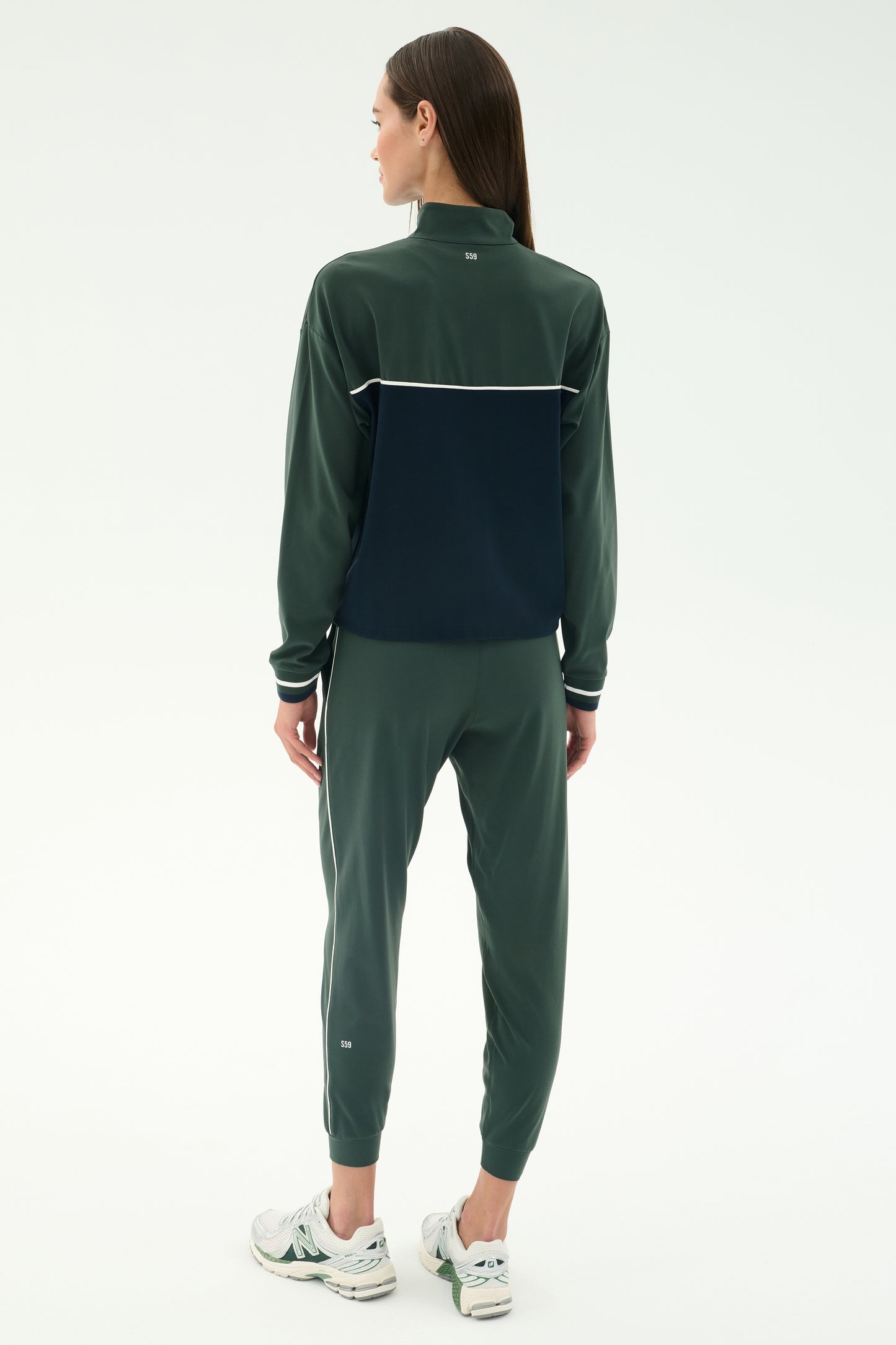 A person faces away from the camera in SPLITS59’s Millie Airweight Half Zip in Military/Indigo, crafted from soft spandex, paired with white sneakers against a plain light background.