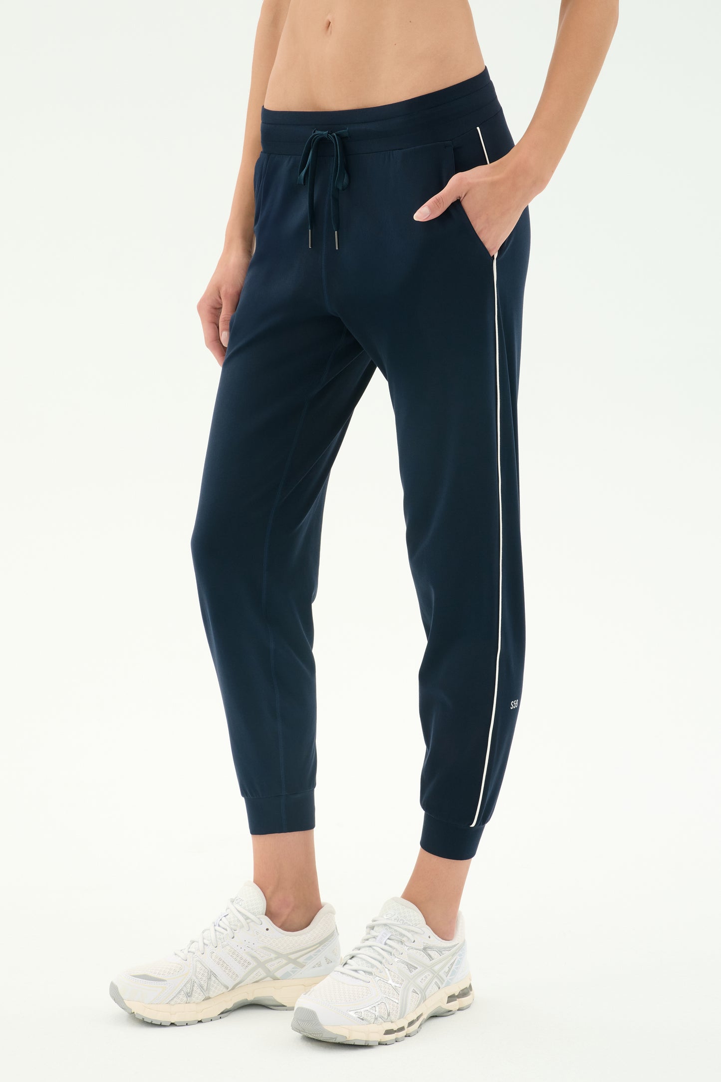 A person wears SPLITS59’s Airweight Jogger 7/8 with Piping in Indigo/White, featuring a relaxed fit, white trim, drawstring waist, and white sneakers. The cropped image shows hands in pockets—ideal for light workouts.