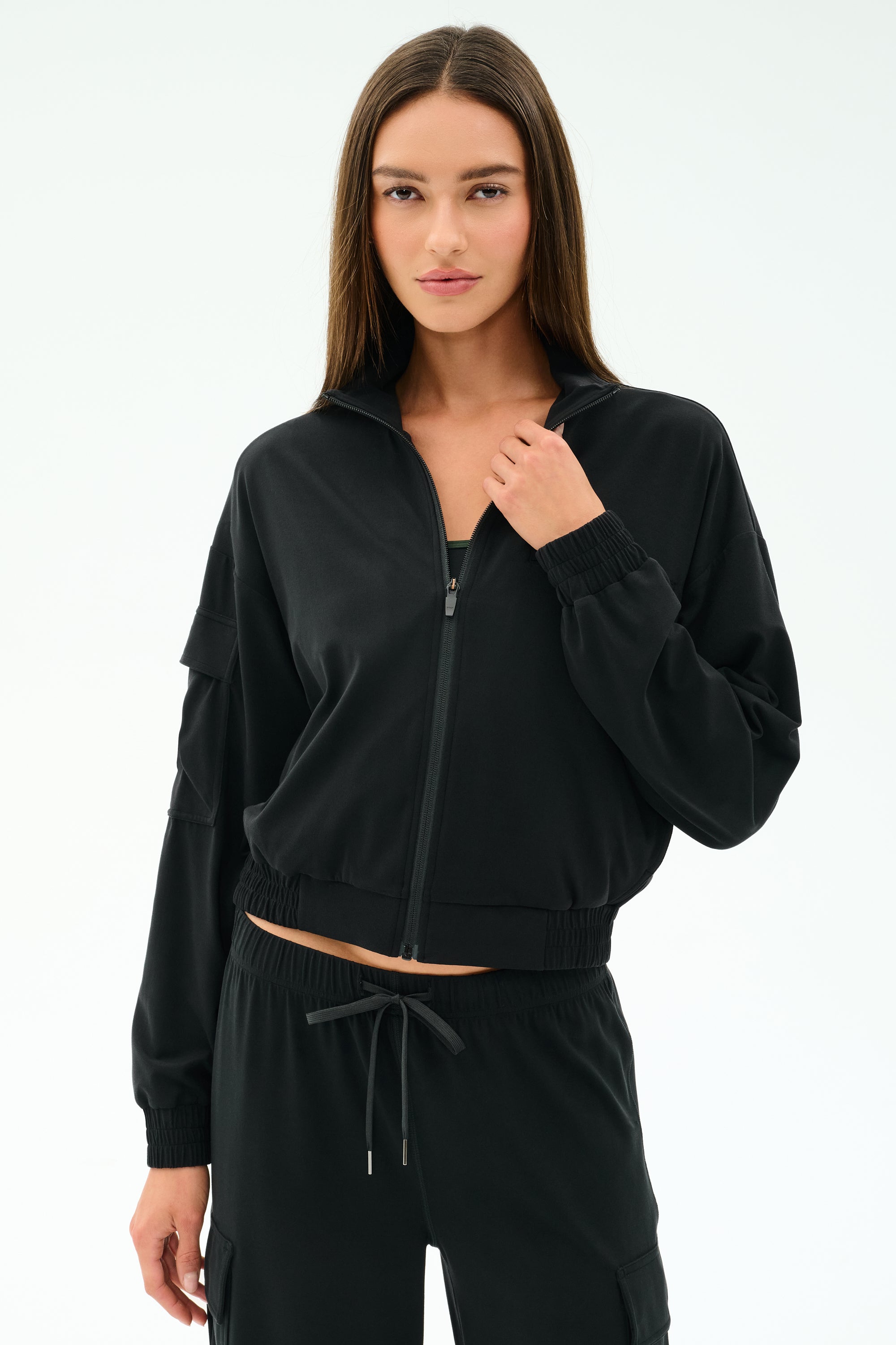 A woman with long brown hair wears the SPLITS59 Peaches Airweight Cargo Jacket in black, paired with matching black stretch pants, standing against a plain white background.