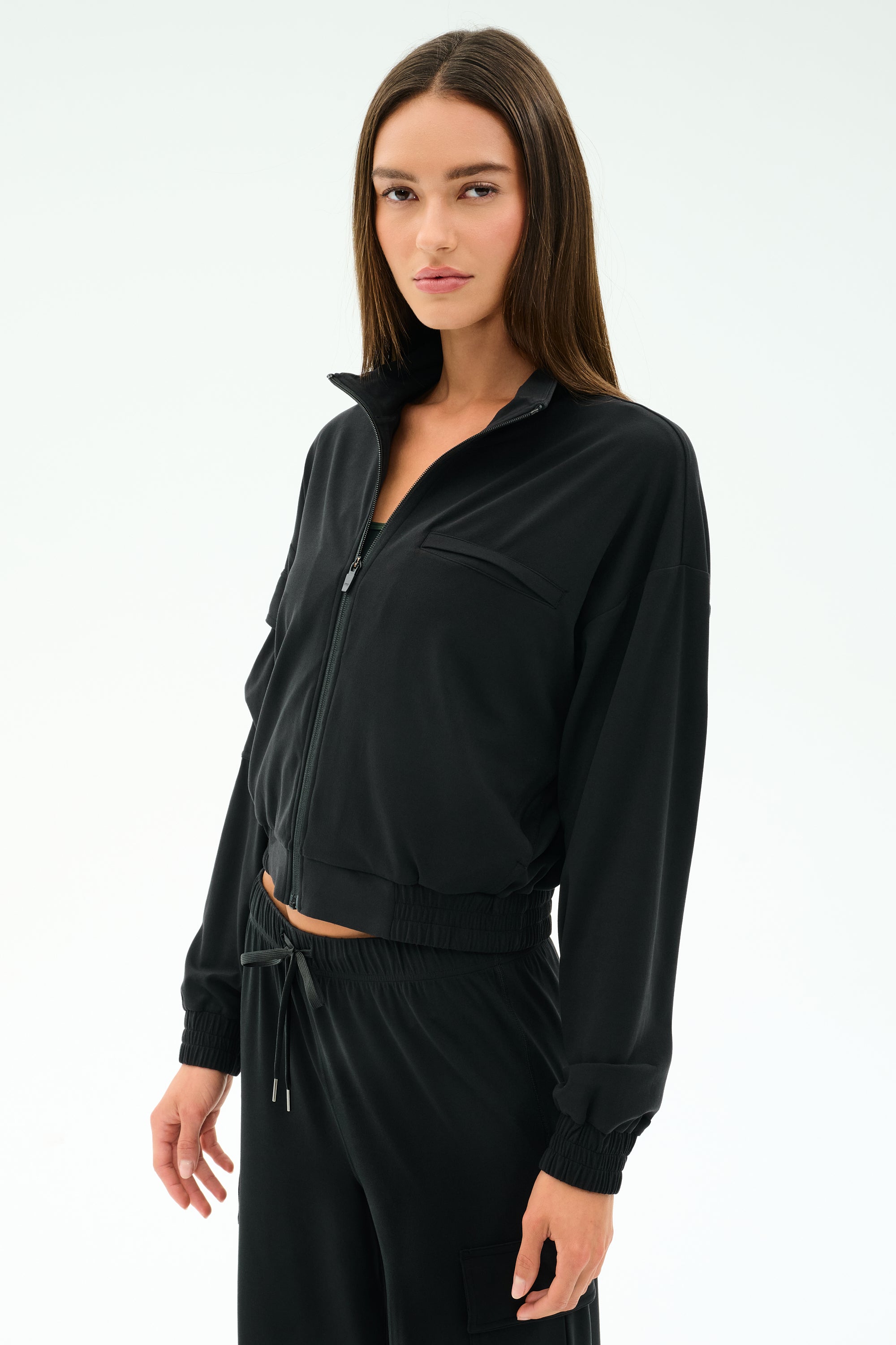 A woman with straight brown hair wears the SPLITS59 Peaches Airweight Cargo Jacket in black, styled with matching black pants, standing against a white background—a sleek, athletic-inspired look.