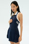A woman wears the SPLITS59 Austin Rigor Dress in Indigo/White, a navy blue sleeveless tennis dress with white trim, standing against a plain light background.