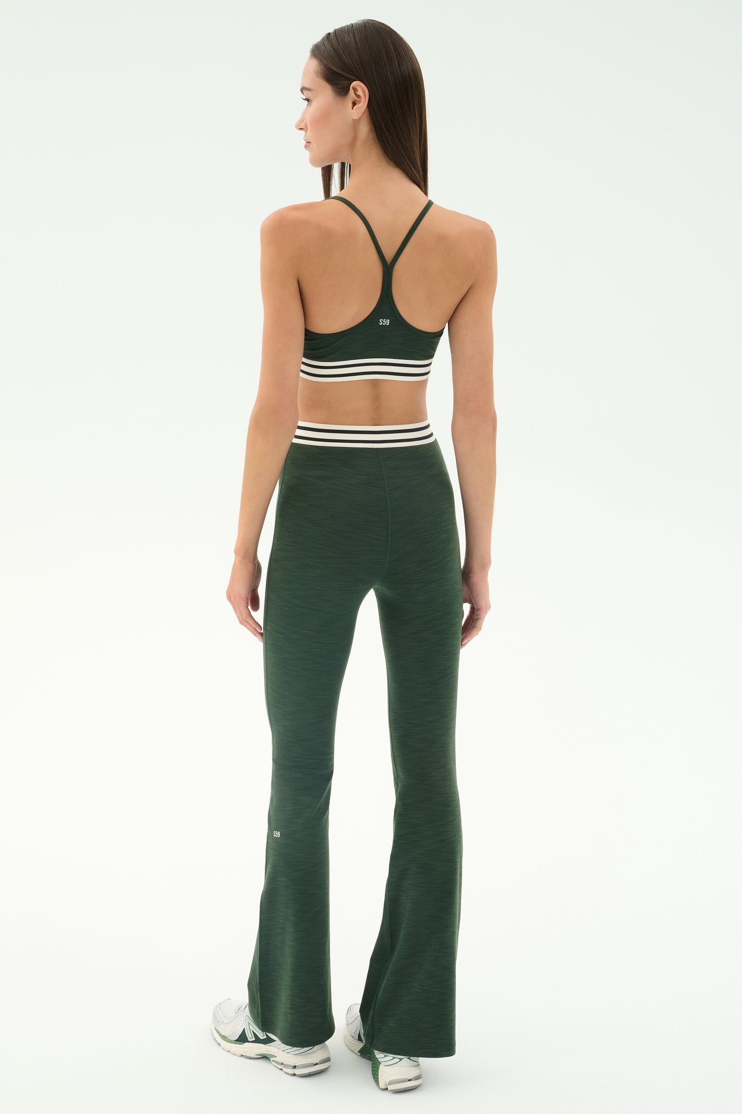 A woman stands facing away in the SPLITS59 Cali Active Rib Bra - Heather Military, paired with matching flared leggings featuring white stripes and white sneakers, set against a plain light background.