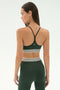 A woman faces away, wearing the SPLITS59 Cali Active Rib Bra in Heather Military—a dark green polyester-spandex sports bra with white stripes—paired with matching high-waisted leggings against a light background.