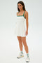 A person stands wearing the SPLITS59 Monah Rigor Dress in White/Arugula, featuring green straps, paired with white sneakers against a plain white background.