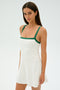 The woman is elegantly dressed in SPLITS59's Monah Rigor Dress in White/Arugula, featuring green spandex straps, against a plain background.
