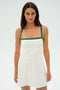 A woman stands against a plain background, wearing the Monah Rigor Dress - White/Arugula by SPLITS59. This sleeveless dress features green straps and elegantly hugs her form, crafted from a blend of spandex and nylon for a comfortable fit.