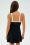 A woman with long brown hair stands facing away in a minimalist setting, wearing the Monah Rigor Dress by SPLITS59. The black sleeveless dress features white straps and is crafted from a spandex blend.