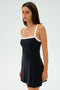 Wearing the SPLITS59 Monah Rigor Dress in black with white straps, a woman stands against a plain background. The dress is crafted from a sleek blend of spandex and nylon as she looks slightly to the side.