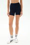 A person wears SPLITS59's Airweight High Waist Short in Indigo, paired with a seamless tank with shelf bra, white socks with black stripes, and white sneakers, standing against a plain light background.
