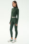 A woman wears the SPLITS59 Bailey Active Rib High Waist 7/8 Legging in Heather Military with a matching green long-sleeve crop top and white sneakers, standing against a plain light background.