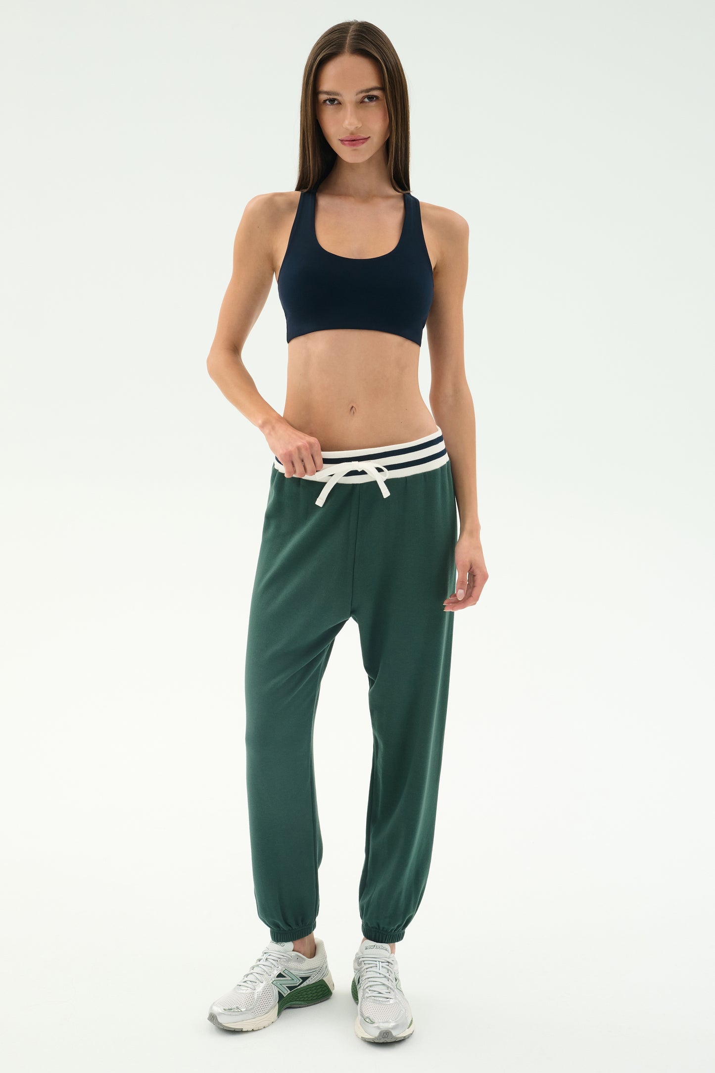 Woman standing against a plain background, wearing a black spandex sports bra, SPLITS59 Andie Oversized Fleece Sweatpant W/ Stripe in Military, and white sneakers.